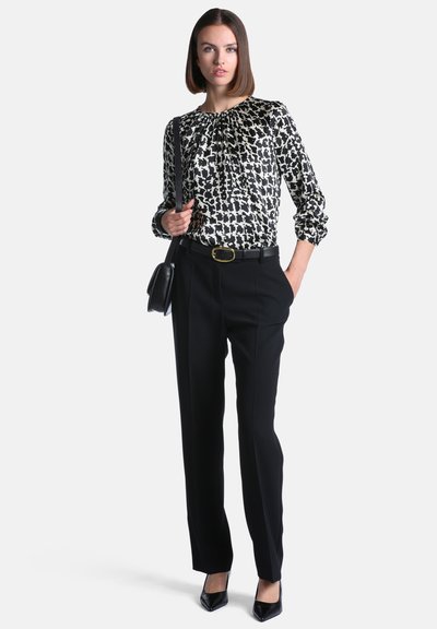 Femme en blouse à motifs noir et blanc, pantalon noir, ceinture noire, talons noirs et sac à main porté à l’épaule, debout avec une main dans la poche.
