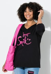 Schwarzes V-Ausschnitt-T-Shirt mit pinkem Grafiketikett "just Smile" und Herzen. Das Model hält eine pinke gepolsterte Tasche und trägt eine Jeans mit Patches.