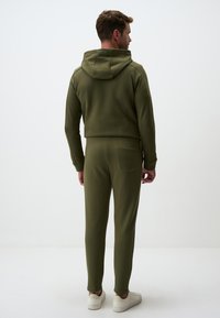 Olivegrüner Hoodie und Jogginganzug aus weichem Material. Der Hoodie verfügt über eine Kordel und eine Kängurutasche; die Jogginghosen haben eine Gesäßtasche.