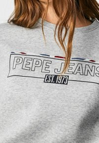 Sudadera gris con texto "PEPE JEANS EST. 1973", llevada por una persona con cabello largo y castaño.