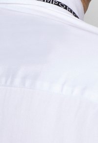 Vit tygskjorta med en enkel design, prydd med ett svart band med texten "Emporio" vid kragen. Slät textur och standardpassform.