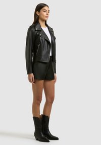 Frau trägt eine schwarze Lederbikerjacke, schwarze Lederhorts, weißes Top und schwarze Cowboy-Stiefel, steht vor einem schlichten Hintergrund.