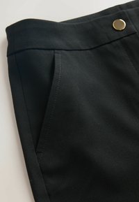 Gros plan sur un pantalon noir avec une poche latérale, un détail à la taille et une fermeture par un bouton doré.
