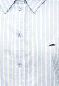 Cămașă cu dungi albastre deschise și albe, cu nasturi și guler ascuțit, având nasturi albastre deschise și un mic logo brodat pe piept.