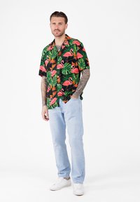 Uomo con tatuaggi che indossa una maglietta nera a maniche corte decorata con fenicotteri rosa e foglie verdi, jeans azzurri e sneakers bianche, in piedi.