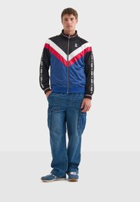 Blå zip-up jacka med röda och vita diagonala ränder, svarta ärmar och logotyp. Kombinerad med löst sittande cargojeans och blå sneakers.