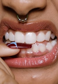 Close-up of a hand holding a long, burgundy nail with a blue design, near glossy white teeth and full lips with a lip gloss finish.