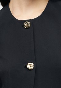 Μαύρο ύφασμα τοπ με στρογγυλή λαιμόκοψη και δύο χρυσά διακοσμητικά κουμπιά κόμπος ως κούμπωμα, με λεία, υφή επιφάνεια.