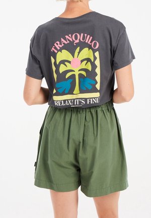 Persona con camiseta gris oscuro con gráfico colorido de palmera y texto "¡Tranquilo Relax! Está bien", combinado con pantalones cortos verdes holgados.