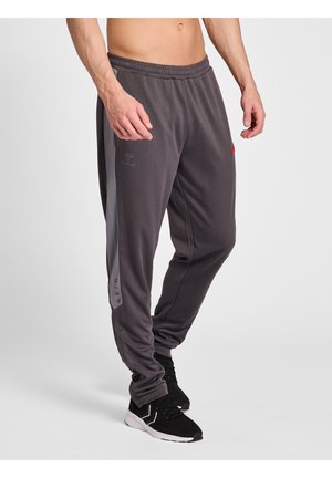 Homme portant un pantalon de sport gris foncé et des chaussures de sport noires, debout devant un fond clair uni, les mains détendues le long du corps.
