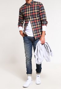 Camisa de cuadros en negro, rojo y amarillo; superpuesta a una camiseta blanca. Lleva unos vaqueros azul y zapatillas blancas. Chaqueta gris en la mano.