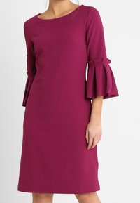 Magenta klänning med trekvartslånga ärmar som har lager av klockärmar. Tyget är slätt med en strukturerad siluett och en rund halsringning.