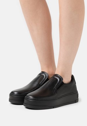 Chaussures compensées noires en cuir, à enfiler, portées pieds nus, avec des bouts lisses et arrondis et des panneaux élastiques sur les côtés pour un enfilage facile.