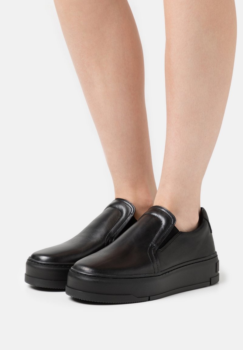 Chaussures compensées noires en cuir, à enfiler, portées pieds nus, avec des bouts lisses et arrondis et des panneaux élastiques sur les côtés pour un enfilage facile.