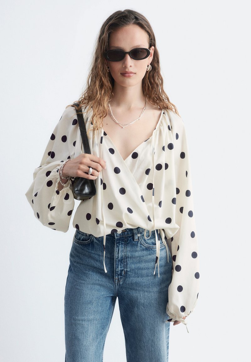 Femme portant des lunettes de soleil noires, un chemisier blanc à pois noirs, un jean bleu et un petit sac à bandoulière noir.