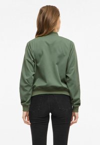 Jachetă bomber verde cu textură netedă, manșete și talie cu tiv, și guler înalt. Este purtată cu pantaloni negri mulați.