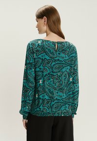 Mujer con cabello castaño lacio que lleva una blusa de manga larga de color teal con un patrón de paisley y pantalones negros, vista desde atrás.