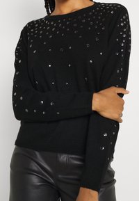 Pull noir à manches avec des clous métalliques ronds sur les épaules, des poignets côtelés et un col rond classique. Tissu doux avec une coupe ajustée.