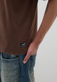 T-shirt di cotone marrone a maniche corte, con un'etichetta laterale. Abbinato a jeans in denim blu chiaro con una texture sbiadita.