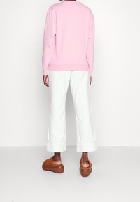 Roze sweatshirt met geribbelde manchetten en zoom, gecombineerd met witte broek met wijde pijpen. Model draagt bruine platformklompen. Minimalistische achtergrond.