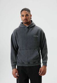 Sweat à capuche gris foncé en tissu doux, avec une poche avant, des poignets côtelés et un logo brodé en blanc. Associé à un pantalon noir.