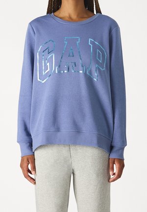 Person iført en blå sweatshirt med skinnende "GAP"-bogstaver og lysegrå bukser, stående mod en ensfarvet hvid baggrund.