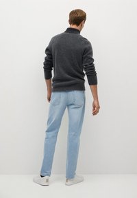 Grauer gestrickter Pullover mit hohem Kragen, kombiniert mit hellblauen Jeans und weißen Sneakern mit goldfarbenen Akzenten. Blick von hinten.