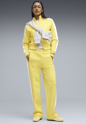 Mujer de pie con las manos en los bolsillos, vistiendo un chándal amarillo y blanco, sudadera gris clara atada alrededor de los hombros y gafas.