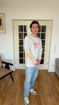 Νεαρός άνδρας στέκεται μέσα σε χώρο με σταυρωμένα τα χέρια, φοράει λευκό πουκάμισο με μακριά μανίκια και σχέδια φλόγας, ανοιχτόχρωμα τζιν και αθλητικά παπούτσια.