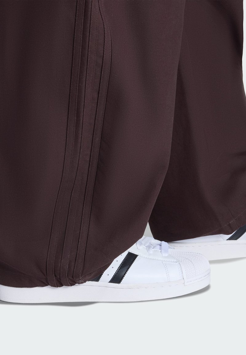 Bordeauxrote Sporthose mit seitlichen Falten, kombiniert mit weißen Turnschuhen mit einem schwarzen Streifen und einer strukturierten Zehenkappe.