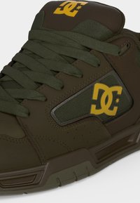 Baskets de la marque DC vert foncé avec des lacets épais et un logo jaune sur le côté et la languette.