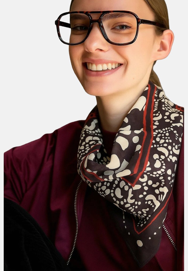 Foulard en soie avec une base marron ornée de motifs organiques blancs et des bords rouges. Le modèle porte de grandes lunettes noires et un haut bordeaux.