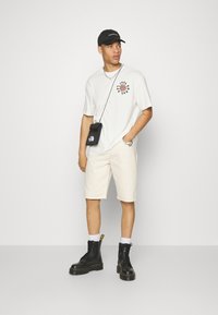Conjunto informal que incluye una camiseta gráfica blanca, pantalones cortos crema, botas de combate negras y un pequeño bolso negro con correa, complementado con una gorra.