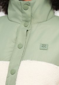 Hele roheline jope, millel on pehme valge fliispaneel, trukkidega nööbid ja väike ruudukujuline logomärgistus rinnal.