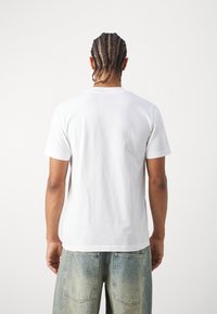 Bílá bavlněná trička s krátkým rukávem, pohled ze zad ukazuje klasický kulatý výstřih a hladkou texturu, v kombinaci se světle modrými džínovými šortkami.