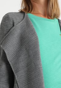 Gråstickad cardigan med texturerade ränder, hängande öppen över en grön T-shirt. Mjuk material, avslappnad design, inga synliga knäppningar.