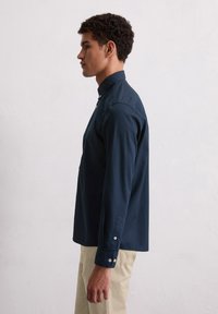 Jonge man in een marineblauwe overhemd met knopen en beige broek staat in profiel tegen een effen lichtgrijze achtergrond.