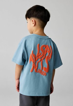 Mladé dieťa s krátkymi tmavými vlasmi, oblečené v svetlomodrom tričku s výraznými oranžovými textúrovanými písmenami na chrbte, stojace pred jednoduchým pozadím.