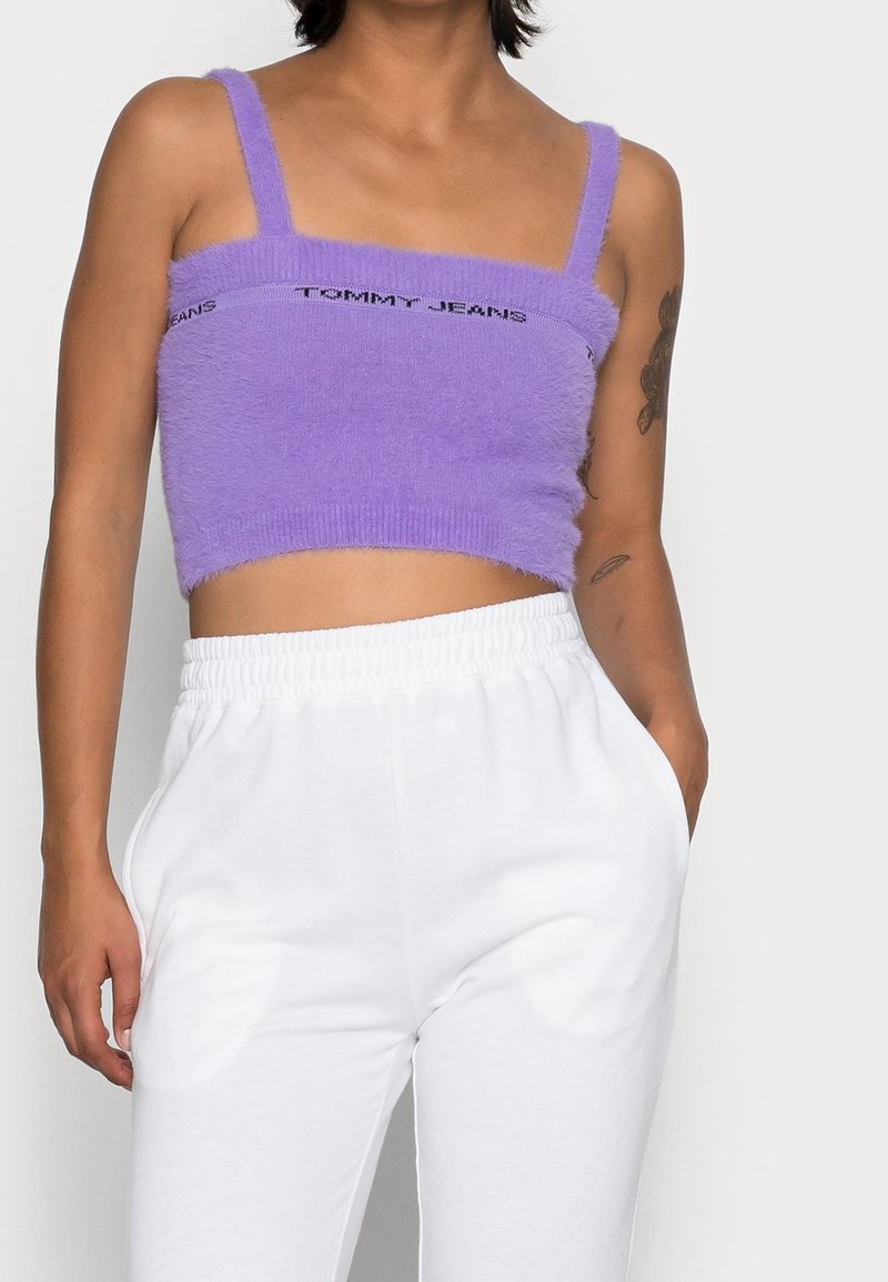 Fioletowy krótki top z teksturowanej tkaniny i regulowanymi ramiączkami, z logo "TOMMY JEANS" na wysokości klatki piersiowej. W połączeniu z białymi spodniami dresowymi.