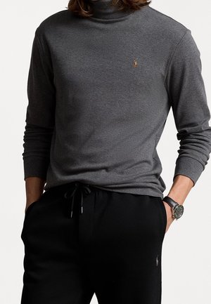 Grauer Rollkragenpullover aus einem weichen Material, mit langen Ärmeln und einem kleinen gestickten Logo auf der Brust. Kombiniert mit schwarzen Hosen.