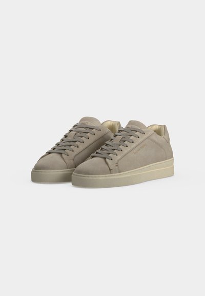 Paire de baskets basses beiges avec lacets, texture lisse et semelles en caoutchouc plates couleur crème sur un fond clair uni.