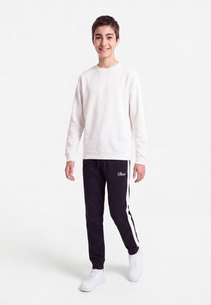 Jeune personne souriante debout, portant un sweat-shirt blanc, un pantalon de survêtement noir avec des bandes blanches et des baskets blanches sur un fond uni.