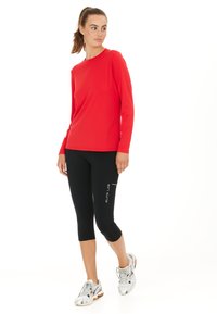 Rotes, langärmliges Sportoberteil mit Rundhalsausschnitt, kombiniert mit schwarzen Leggings bis zur Mitte der Wade. Model trägt silberne Laufschuhe mit Mustern.