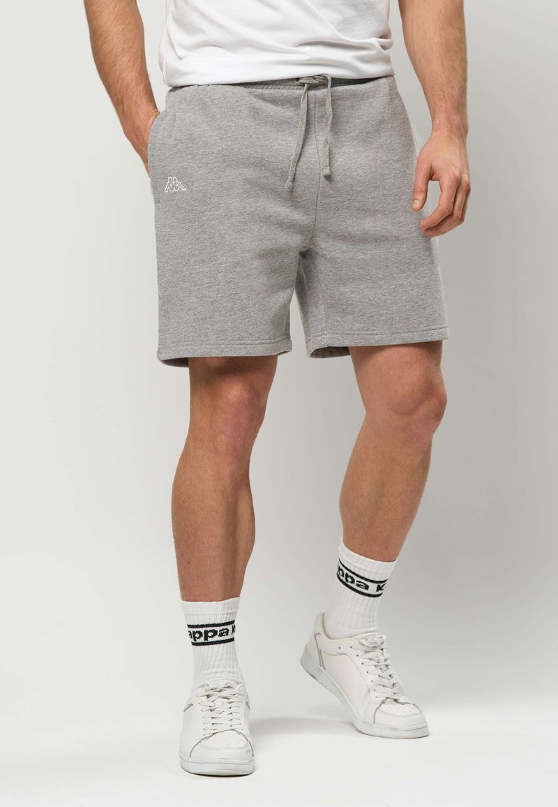 Muškarac u svijetlosivim kratkim hlačama s vezicama, bijelim čarapama s crnim logotipom i bijelim tenisicama, stoji s jednom rukom u džepu.