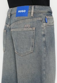 Világoskék mosású farmernadrág, nagy kék "HUGO" feliratos címkével, két hátsó zsebbel és finom varrási részletekkel.