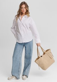 Chemise blanche à boutons, jeans ample bleu clair, sac fourre-tout en suède beige avec des anses, et baskets avec des accents beiges et gris.
