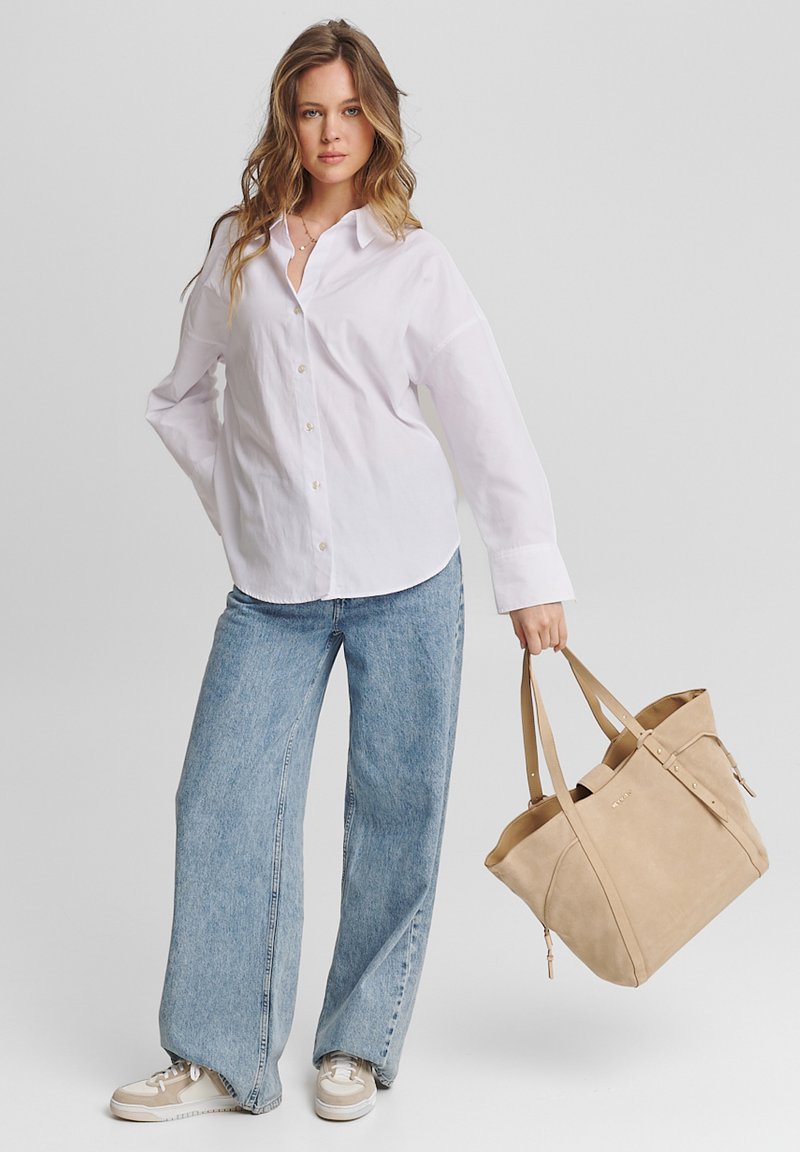 Chemise blanche à boutons, jeans ample bleu clair, sac fourre-tout en suède beige avec des anses, et baskets avec des accents beiges et gris.