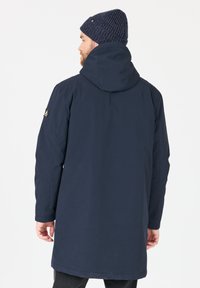 Parka impermeable de color azul marino con capucha, que presenta una textura suave y un parche con el logotipo en la manga izquierda. Llevada con un gorro de lana.