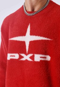 Rode fuzzy trui met een ontwerp van een witte ster en de letters "PXP". Gemaakt van een zachte stof met een gestructureerde afwerking. Kettinghalsketting zichtbaar.