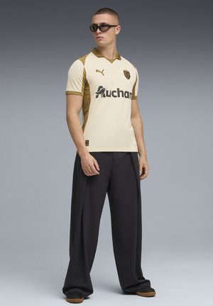 Beige Sporttrikot mit goldenen Akzenten, kurzen Ärmeln und schwarzer Markenkennzeichnung. Lockere schwarze Hosen und dunkle Sneakers vervollständigen das Outfit.